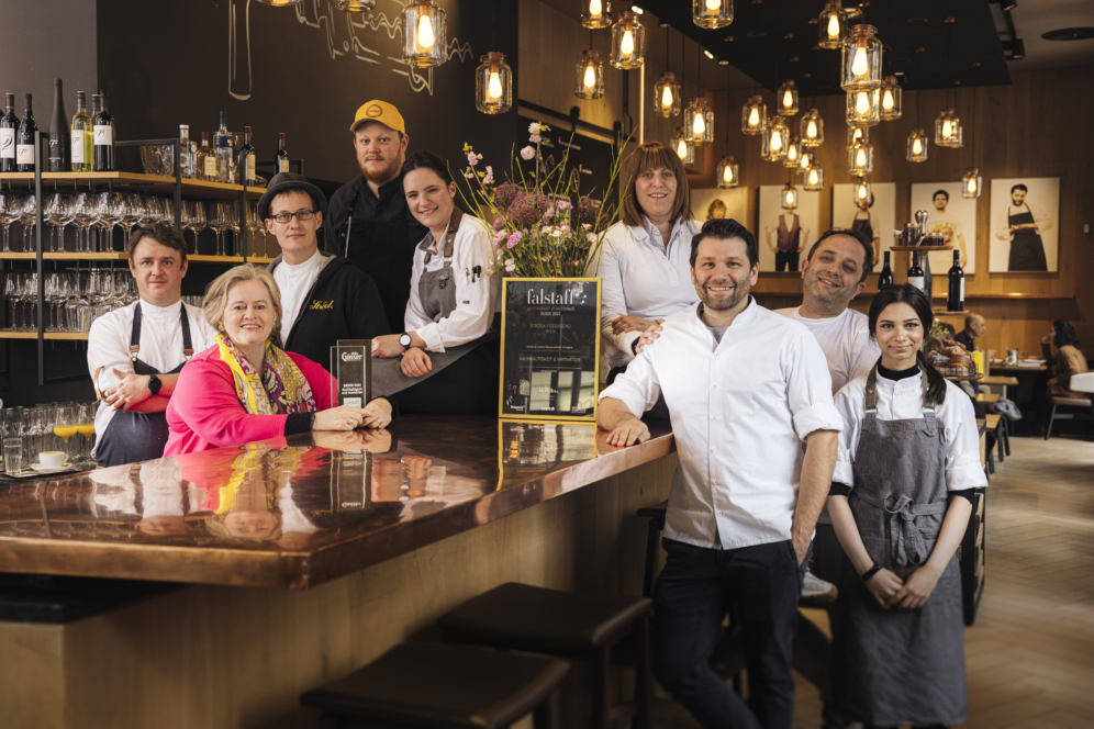 Eine Gruppe von Köchen und Restaurantmitarbeitern posiert lächelnd hinter einer polierten Bar in einem warm beleuchteten, modernen Restaurant mit Hängelampen und Regalen mit Flaschen und Gläsern im Hintergrund.