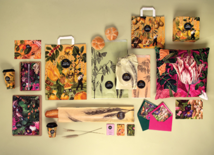 Eine Vielzahl von bunten Verpackungen, Umschlägen, Kaffeetassen und Tüten mit lebhaften botanischen und floralen Illustrationen sind auf einer hellen Fläche angeordnet, zusammen mit Brot, Gebäck und Markenaufklebern.