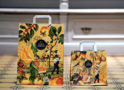 Zwei bunte Papiertüten mit Obst- und Pflanzenmotiven stehen auf einem gemusterten Kachelboden. Jede Tüte zeigt eine kleine Figur, die einen Wagen schiebt, und auf der Vorderseite die Aufschrift "Feierabend" in einem schwarzen Kreis.