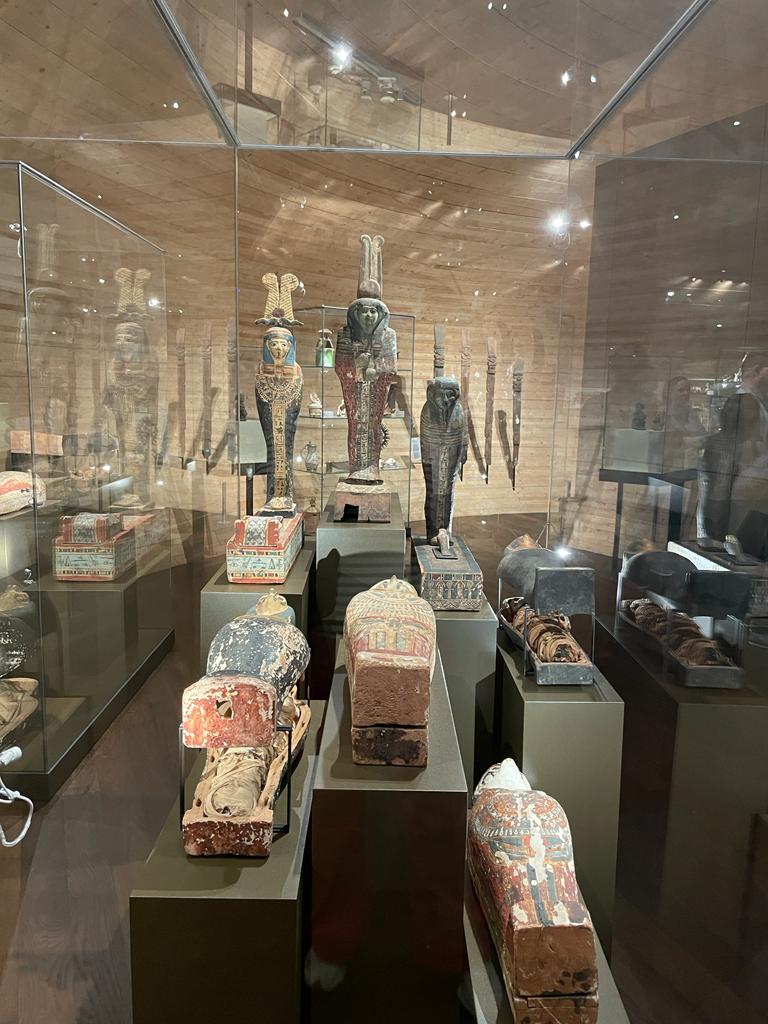 Altägyptische Artefakte in einer Museumsvitrine mit mehreren bemalten Holzsarkophagen und Statuen, darunter auch tierköpfige Figuren, an einer Holzwand mit Spiegelungen auf dem Glas.