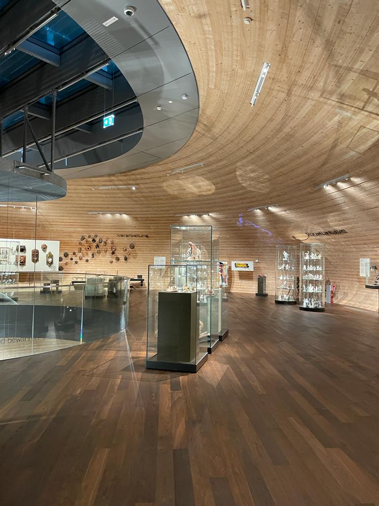 Eine moderne Museumsgalerie mit geschwungenen Holzwänden und -decken, Holzböden und Glasvitrinen, in denen verschiedene Artefakte ausgestellt sind. Eine sanfte Beleuchtung schafft eine ruhige Atmosphäre, und die Objekte sind an den Wänden und in Vitrinen angebracht.