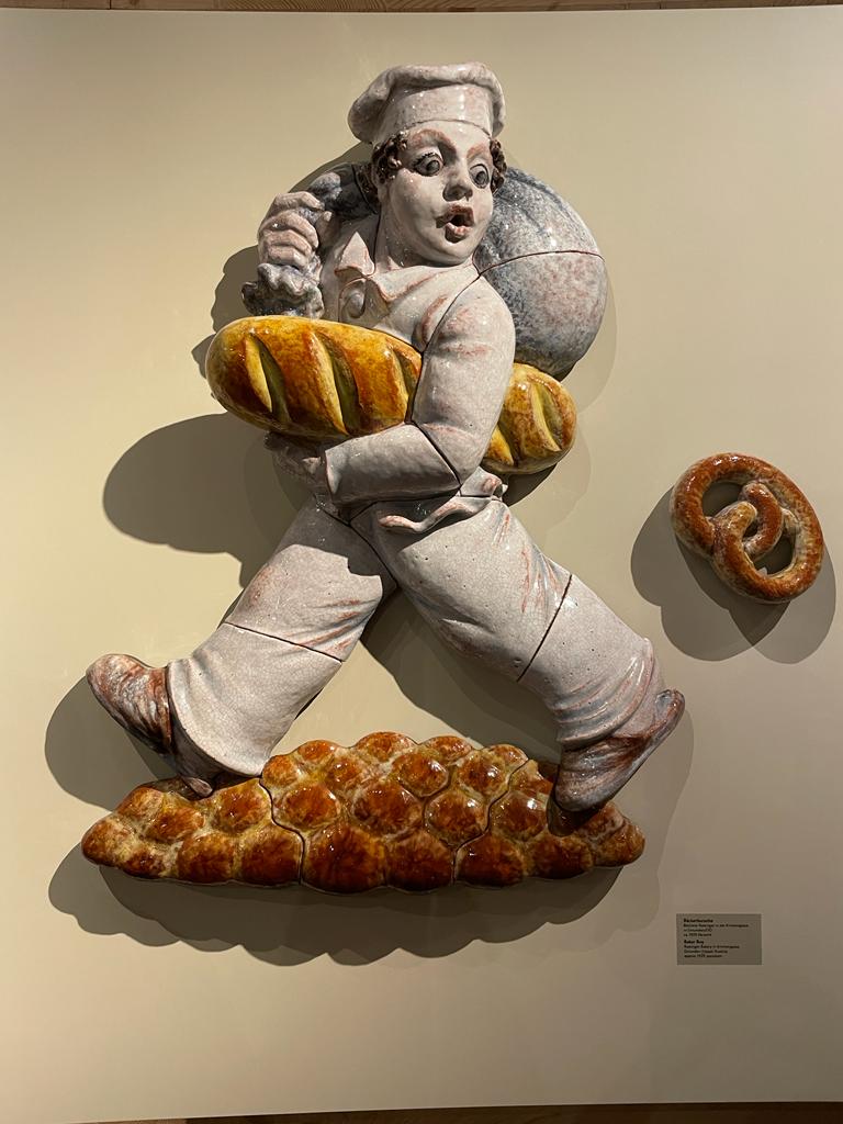 Keramische Wandskulptur eines Bäckers in weißer Kleidung, der einen großen Laib Brot trägt, daneben eine Brezel und Brötchen, alles auf einem hellen Hintergrund.