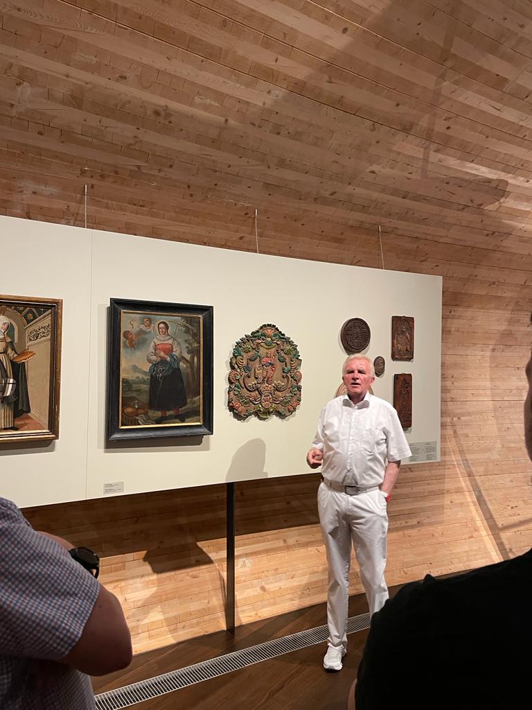 Ein ganz in Weiß gekleideter Mann steht und spricht vor einer Galeriewand, die verschiedene Kunstwerke und Artefakte in einem Raum mit Holzwänden und einer gewölbten Decke zeigt. Im Vordergrund sind mehrere Personen zu sehen.
