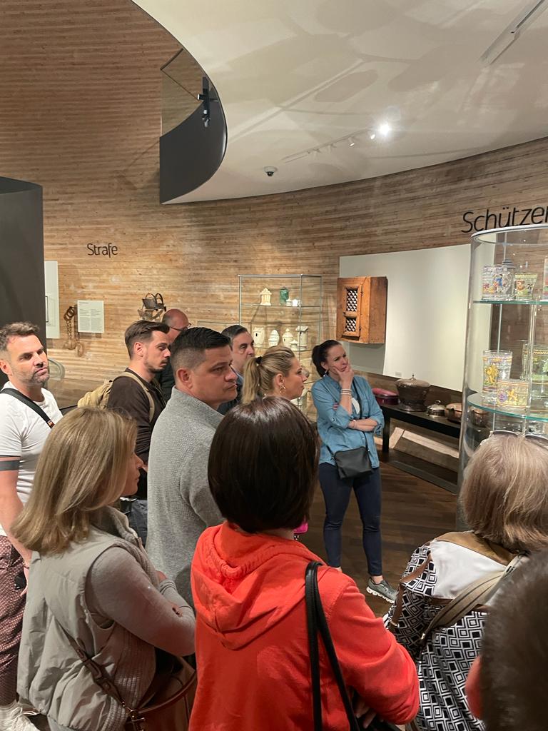 Eine Gruppe von Menschen steht dicht beieinander, hört aufmerksam zu und betrachtet die Ausstellungsstücke in einem Museum mit Holzwänden und Glasvitrinen, die Artefakte enthalten.