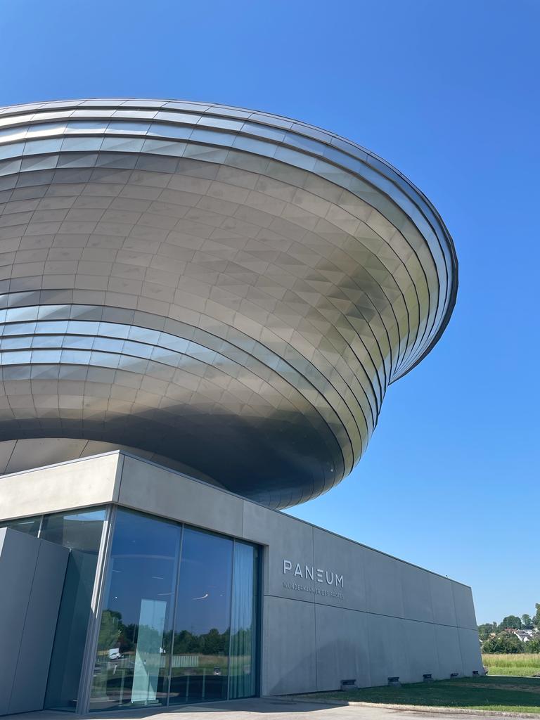 Ein modernes Gebäude mit einer großen, ovalen Metallstruktur über einem Eingang aus Glas und Beton. Auf dem Gebäude ist der Schriftzug "PANEUM" vor einem strahlend blauen Himmel zu sehen.