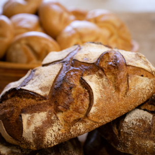 Nahaufnahme von rustikalen, knusprigen, runden Brotlaiben mit goldbrauner, strukturierter Oberfläche und dekorativen Einkerbungen. Im Hintergrund befindet sich ein Korb mit mehreren kleineren Brötchen.