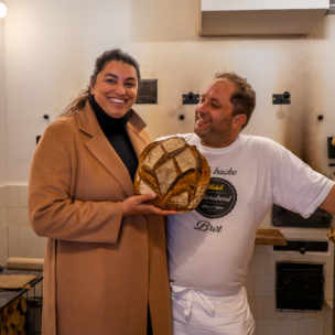 Eine lächelnde Frau in einem braunen Mantel hält einen großen runden Laib Brot in der Hand. Sie steht neben einem Mann in einem weißen Hemd und einer Schürze in einer Bäckerei mit weiß gekachelten Wänden und Backgeräten.
