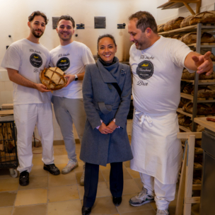 Vier Personen posieren in einer Bäckerei, drei Männer in weißen Uniformen mit Brot und eine Frau in einem grauen Mantel. Regale mit frischen Broten füllen den Hintergrund, und die Gruppe wirkt fröhlich und freundlich.