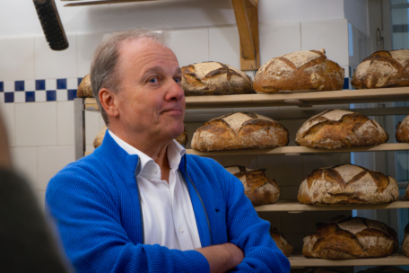 Ein Mann in einem blauen Pullover steht mit verschränkten Armen in einer Bäckerei, hinter ihm stapeln sich große, knusprige Brote in den Regalen.