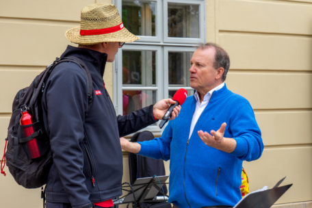Zwei Männer unterhalten sich draußen; der eine mit Strohhut und schwarzer Jacke, mit Rucksack und roter Wasserflasche, der andere, der einen blauen Pullover trägt, hält ein rotes Mikrofon. Sie stehen vor einer beigen Wand und einem Fenster.