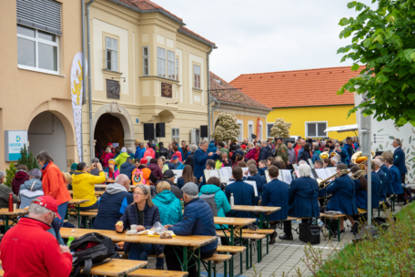 Eine große Gruppe von Menschen, darunter eine Blaskapelle, sitzt auf Holzbänken bei einer Veranstaltung im Freien auf einem Stadtplatz mit historischen Gebäuden im Hintergrund. Viele tragen bunte Jacken und sind in ein Gespräch vertieft.