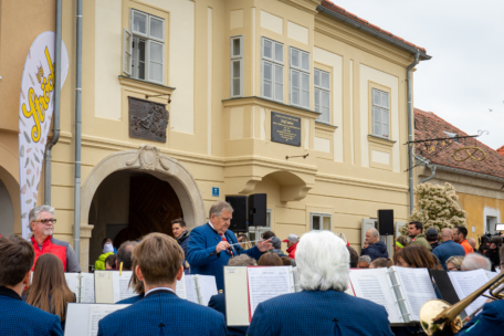 Ein Dirigent leitet eine Kapelle, die im Freien vor einem historischen gelben Gebäude spielt, während eine Menschenmenge zusieht. Die Musiker tragen blaue Jacken, und auf der linken Seite ist ein Schild mit der Aufschrift Svečanost zu sehen.