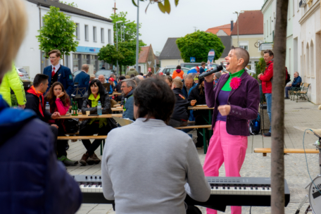 Eine lächelnde Frau in leuchtend rosa Hosen und einer lila Jacke singt in ein Mikrofon, begleitet von einem Keyboarder, während die Menschen an Tischen im Freien sitzen und die Live-Musik auf einem belebten Stadtplatz genießen.
