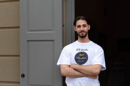Ein junger Mann mit kurzen dunklen Haaren und einem Bart steht mit verschränkten Armen vor einer teilweise geöffneten grauen Tür und trägt ein weißes T-Shirt mit einem kreisförmigen Logo und Text. Der Hintergrund ist neutral und in Innenräumen.