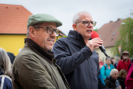 Zwei Männer mit Brillen sprechen bei einer Veranstaltung im Freien; einer hält ein rotes Mikrofon. Im Hintergrund sind Menschen versammelt, man sieht Häuser und einen bewölkten Himmel.