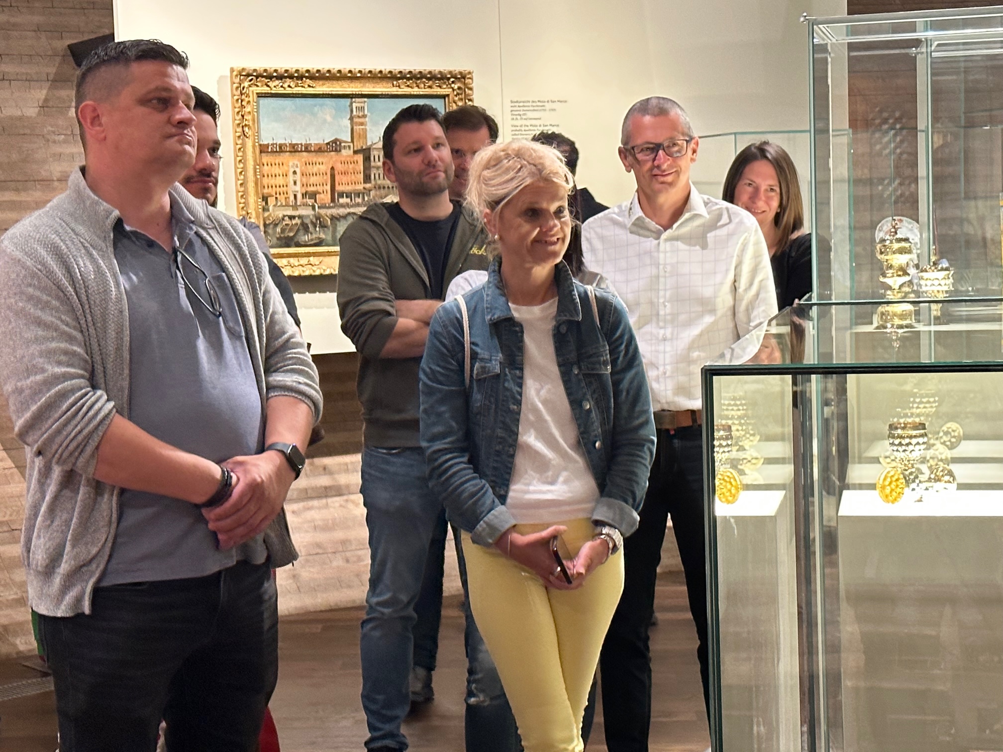 Eine Gruppe von Menschen steht aufmerksam in einem Museum und betrachtet Glasvitrinen mit Artefakten. Hinter ihnen hängt ein gerahmtes Gemälde an der Wand, und alle scheinen konzentriert und engagiert zu sein.