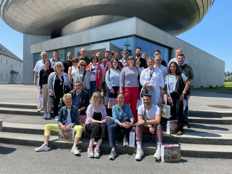Eine Gruppe von etwa 30 Personen posiert für ein Foto auf einer Freitreppe vor einem modernen Gebäude mit einem metallischen, geschwungenen Design unter blauem Himmel. Viele lächeln und halten Taschen in der Hand.