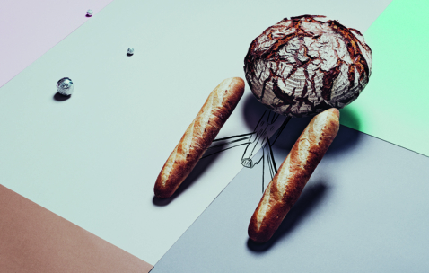 Ein runder Laib Bauernbrot und zwei Baguettes sind auf einem pastellfarbenen geometrischen Hintergrund angeordnet, der an ein Tier mit gezeichneten Beinen und einem Schwanz darunter erinnert. Kleine Metallkugeln sind in der Nähe verstreut.