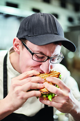 Eine Person mit Brille, schwarzer Mütze und weißem Kochkittel nimmt in einer Küche einen großen Bissen von einem mit Salat und anderen Zutaten belegten Sandwich.
