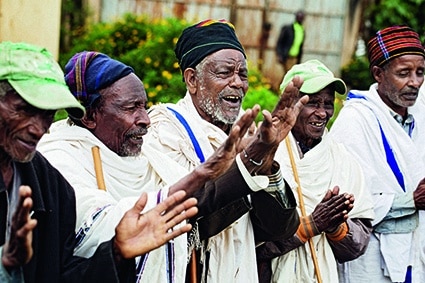 Eine Gruppe älterer Männer in traditioneller Kleidung und mit Hüten steht im Freien nebeneinander, klatscht und lächelt und scheint an einem fröhlichen Gemeinschaftsereignis teilzunehmen.