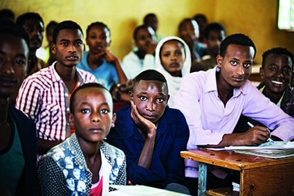 Eine Gruppe von Schülern, zumeist Teenager, sitzt in einem Klassenzimmer dicht an den Tischen und blickt aufmerksam nach vorne. Das Klassenzimmer hat gelbe Wände und natürliches Licht, das durch ein Fenster einfällt.