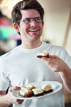 Ein lächelnder Mann mit Brille und weißem T-Shirt hält einen Teller mit Essen in einer Hand und hebt mit der anderen ein Stück zum Mund. Der Teller enthält mehrere kleine Stücke, die mit einem cremigen Aufstrich belegt sind.