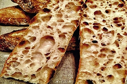 Zwei Scheiben rustikales Brot mit einer dunklen, knusprigen Kruste und einer offenen, luftigen Krumenstruktur voller großer Löcher, die auf einer leichten Stoffoberfläche ruhen.