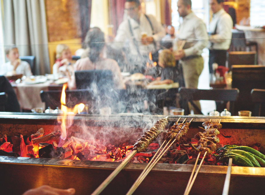 Fleisch- und Gemüsespieße, die in einem Restaurant über heißen Kohlen gegrillt werden, während im Hintergrund Rauch aufsteigt und die Gäste an den Tischen sitzen, während sich die Kellner um die Gäste kümmern.