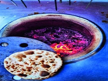Nahaufnahme eines Tandoor-Ofens mit glühender Holzkohle, in dem ein Stück Fladenbrot gebacken wird, das von einem Metallwerkzeug in der Nähe der Ofenöffnung gehalten wird.