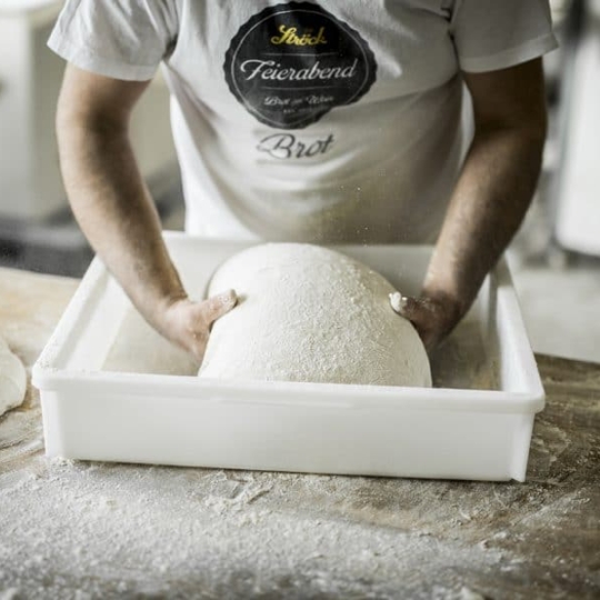 Eine Person knetet eine große Teigkugel in einem weißen Plastikbehälter auf einer bemehlten Holzfläche und trägt ein weißes T-Shirt mit dem Logo einer Bäckerei.