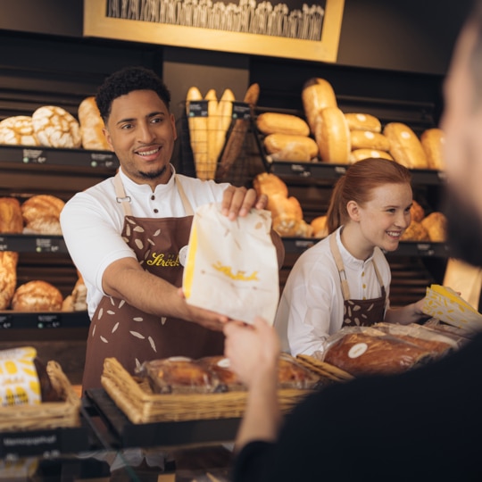 Eine lächelnde Bäckereiangestellte reicht einem Kunden über den Tresen hinweg eine Papiertüte, während ein anderer Angestellter in der Nähe steht; in den Regalen hinter ihnen liegen verschiedene Brote.
