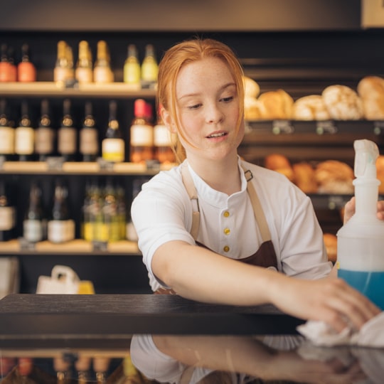 Eine junge Frau mit roten Haaren und einer Schürze wischt in einer Bäckerei eine Theke mit einem Tuch und Reinigungsspray ab, während im Hintergrund Regale mit Brot und Flaschen zu sehen sind.