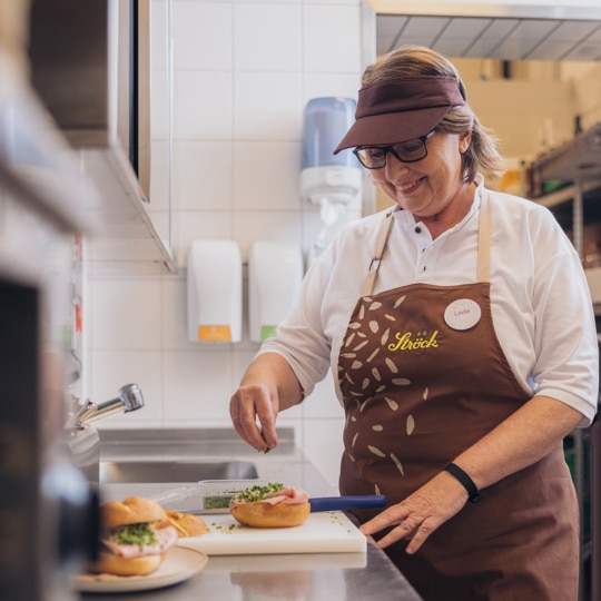 Eine Frau mit Brille, Visier und brauner Schürze bereitet in einer hellen, sauberen Küche Sandwiches zu und lächelt, während sie die Zutaten zu den Speisen auf der Theke hinzufügt.