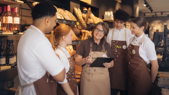 Ein lächelnder Manager mit einem Klemmbrett spricht mit vier Bäckereimitarbeitern in braunen Schürzen, die gemeinsam vor Regalen mit Brot und Weinflaschen stehen. Die Gruppe macht einen engagierten und aufmerksamen Eindruck.