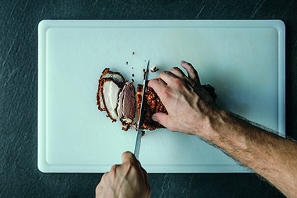 Eine Person schneidet mit einem scharfen Messer ein Stück gekochtes Fleisch auf einem weißen Schneidebrett auf, wobei eine Hand das Fleisch hält und die andere Hand schneidet. Der Hintergrund ist eine dunkle Arbeitsplatte.