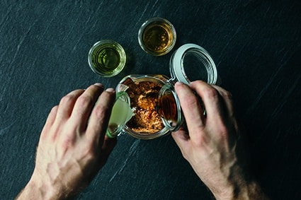 Eine Person gießt eine Flüssigkeit aus einem kleinen Glas in ein Glas mit einer braunen Mischung, während zwei weitere kleine Gläser mit Flüssigkeiten auf einer dunklen Fläche in der Nähe stehen.