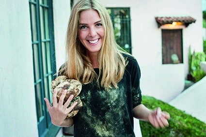 Eine lächelnde Frau mit langen blonden Haaren steht draußen und hält eine große, unregelmäßig geformte Trüffel in einer Hand. Sie trägt ein schwarzes Hemd mit Schmutz- oder Staubflecken darauf.