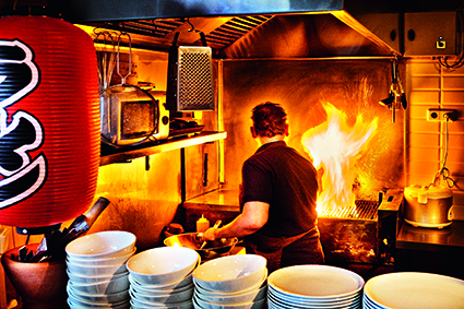 Ein Chefkoch kocht in einer geschäftigen Restaurantküche mit großen Flammen auf dem Herd. Im Vordergrund stapeln sich weiße Schüsseln, und auf der linken Seite hängt eine rote Laterne mit japanischer Schrift. Die Szene ist lebendig und pulsierend.