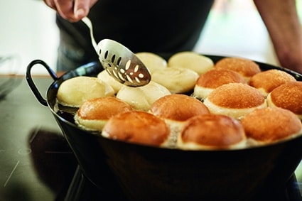 Eine Person benutzt einen Schaumlöffel, um in heißem Öl frittierte Krapfen zu wenden, wobei einige Krapfen goldbraun und andere noch blass sind.