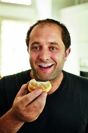 Ein lächelnder Mann mit kurzen Haaren hält einen gepuderten Donut mit Puderzucker auf Mund und Lippen. Er trägt ein dunkles Hemd und steht drinnen in einem hell erleuchteten Raum.