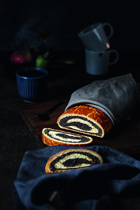 Ein aufgeschnittener Laib Strudelbrot liegt teilweise in ein Tuch eingewickelt auf einem Holzbrett, mit zwei Scheiben im Vordergrund und Bechern, einer Tasse und Früchten, die im dunklen Hintergrund verschwimmen.