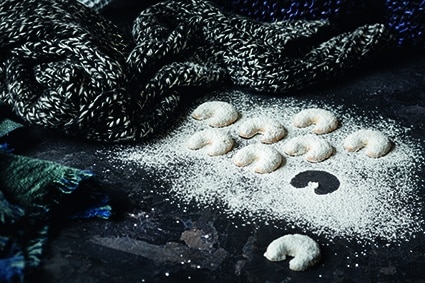 Auf einer dunklen, mit Puderzucker bestäubten Fläche sind halbmondförmige Kekse angeordnet, daneben ein schwarz-weißer Strickschal und ein grün-blaues gewebtes Tuch.