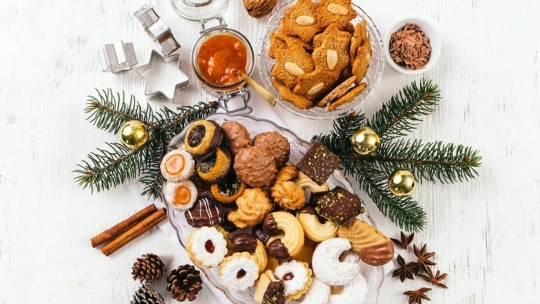 Eine festliche Platte mit verschiedenen Weihnachtsplätzchen und Leckereien, umgeben von Tannenzweigen, goldenen Ornamenten, Tannenzapfen, Sternanis, Zimtstangen und Ausstechformen auf einer weißen Holzoberfläche.