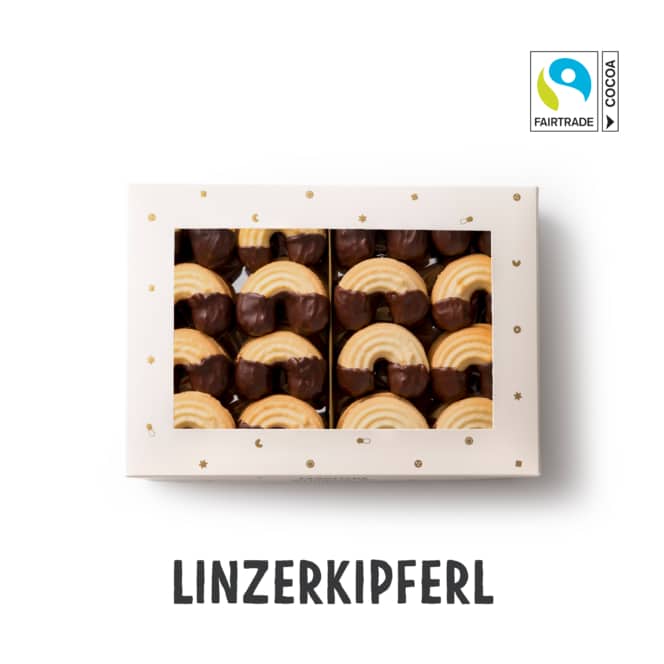 Eine Schachtel mit Linzerkipferl, halb in Schokolade getaucht, fein säuberlich in Reihen angeordnet. Die Schachtel hat ein durchsichtiges Fenster und ist mit goldenen Sternen verziert. In der rechten oberen Ecke ist ein Etikett mit Fairtrade-Kakao angebracht.