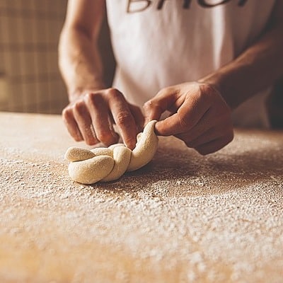 Nahaufnahme der Hände einer Person, die auf einer bemehlten Holzfläche Teig flechtet und Brot zubereitet. Die Person trägt ein weißes Hemd, und der Arbeitsbereich ist schwach beleuchtet.