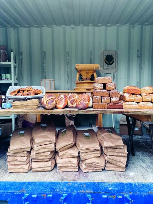 Ein Marktstand bietet frisches Brot, Baguette und Gebäck auf Holztischen an, unter denen sich große braune Papiertüten mit Mehl stapeln. Im Hintergrund sind eine Getreidemühle und Regale zu sehen.