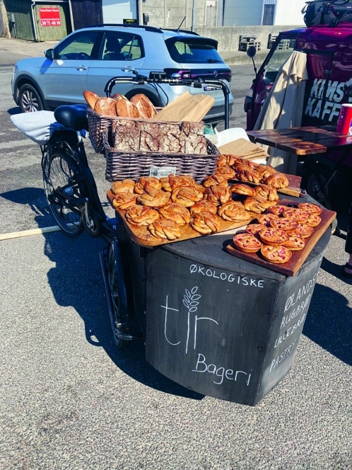 Ein Fahrradkarren stellt Körbe mit verschiedenen Brotsorten und Gebäck in einer sonnigen Straße aus. Auf einem Kreideschild an dem Wagen steht tlr Bageri und Økologiske. Im Hintergrund sind ein Auto und ein Lieferwagen geparkt.