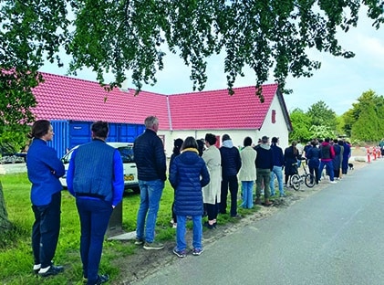 Eine Gruppe von Menschen steht in einer langen Reihe auf einem Bürgersteig neben einem weißen Gebäude mit rotem Dach, einige tragen Jacken und schauen von der Kamera weg unter einen großen, belaubten Baum.