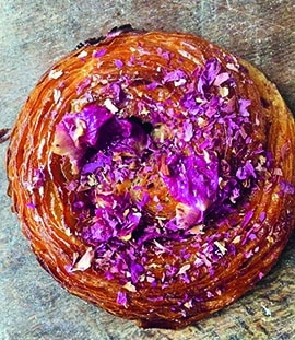 Auf einer rustikalen Holzunterlage liegt ein runder Blätterteig, der mit leuchtend violetten Blütenblättern belegt ist.