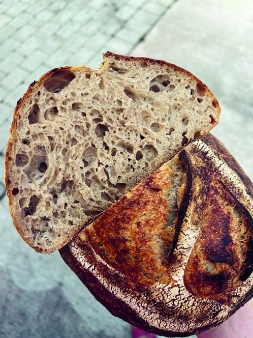 Nahaufnahme eines rustikalen Sauerteigbrotes, bei dem die eine Hälfte aufgeschnitten ist und die feuchte, luftige Krume zum Vorschein kommt, während die andere Hälfte eine dunkle, knusprige Außenseite zeigt, mit einem verschwommenen Steinpflaster im Hintergrund.
